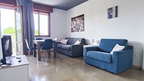 A modern living room with a blue armchair, grey sofa, and a dining area with a table and chairs. - Cozy Studio With Balcony (Jesolo)
