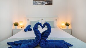 Una cama con una toalla dispuesta en forma de corazón, un cuadro de un paisaje costero y dos lámparas de noche.
