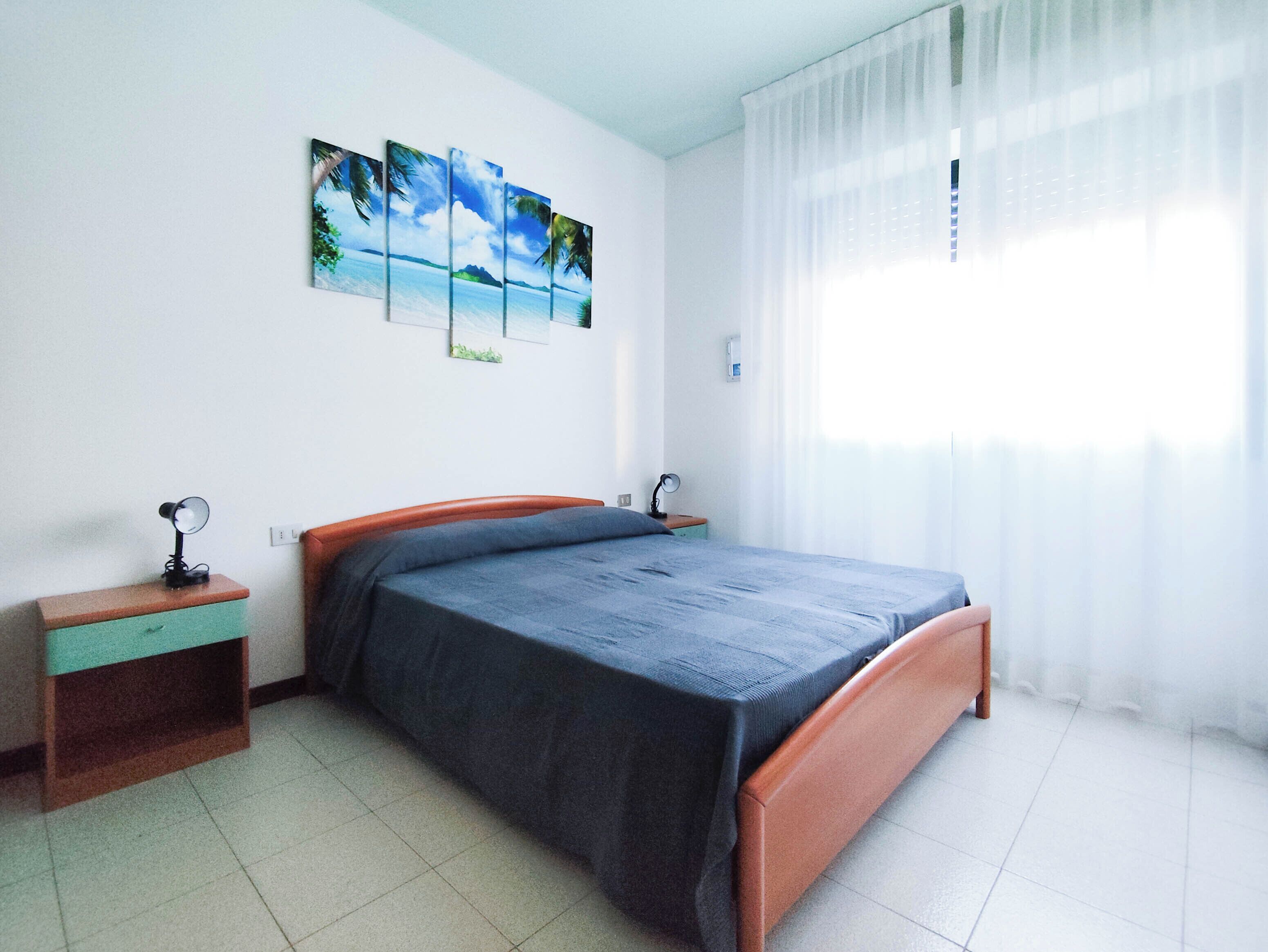 A bedroom with a bed, two bedside tables, a nightstand, a window with sheer curtains, and a wall-mounted artwork depicting a tropical scene.