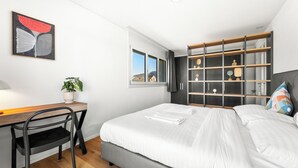 A modern bedroom with a bed, a desk, a chair, a plant, a window, and a built-in shelving unit. - Appartement B3 13 (Avenches)