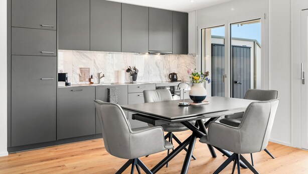 A modern kitchen with grey cabinets, a marble backsplash, and a dining area with a table and chairs. - Appartement B3 13 (Avenches)