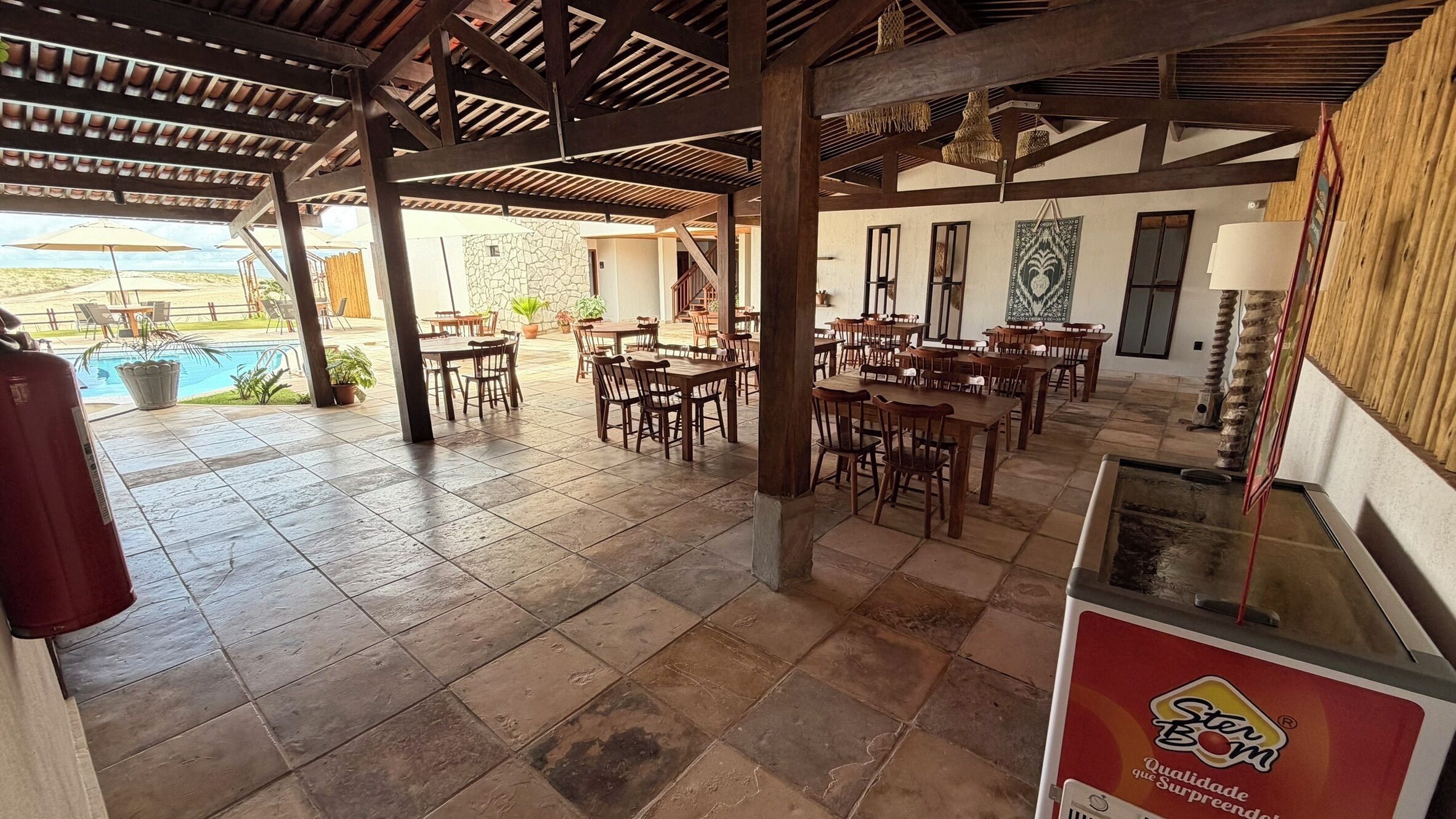 Un espacio comedor al aire libre cubierto con baldosas de piedra, mesas y sillas de madera, y vista a una piscina.