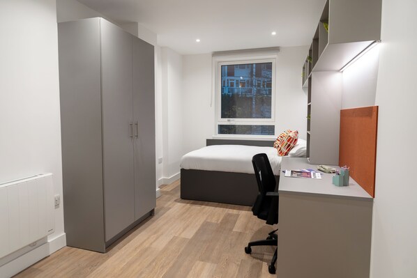 Un dormitorio moderno con un armario grande, una cama con cabecera gris, un escritorio con una silla y una ventana con vistas a edificios.