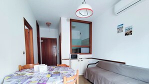 A compact living space with a dining table, chairs, a sofa, a kitchenette, and a door leading to another room. - Seaside Apartment With Balcony (Jesolo)