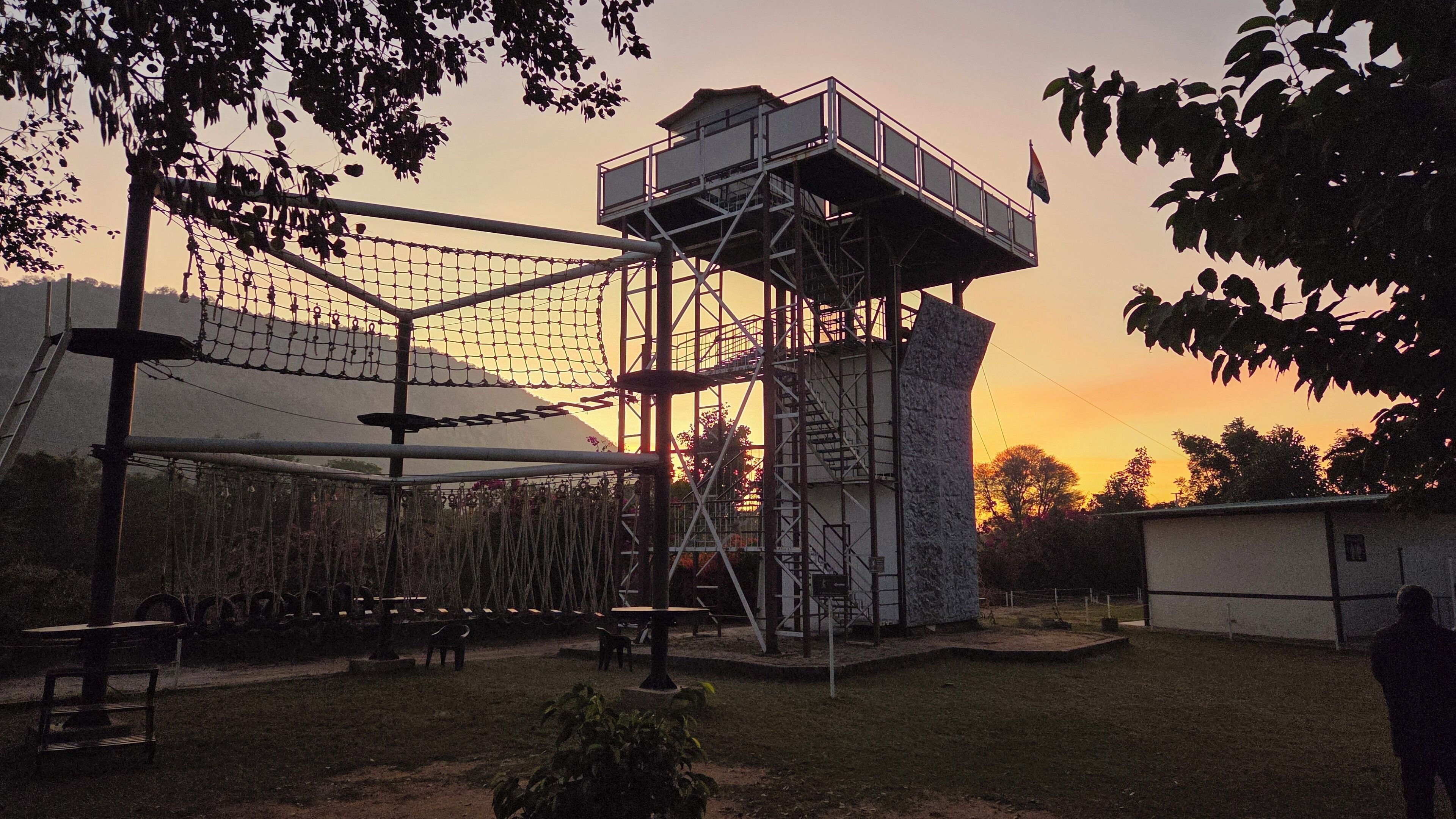 A netted climbing structure with a viewing platform, surrounded by trees and a small building.