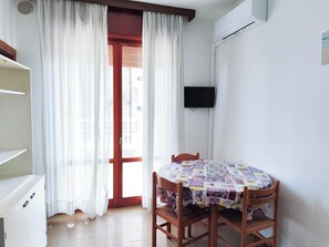 Ein Zimmer mit einem Esstisch, bedeckt mit einer blumigen Tischdecke, zwei Holzsesseln, einem an der Wand montierten Fernseher und einer Glastür mit weißen Vorhängen.