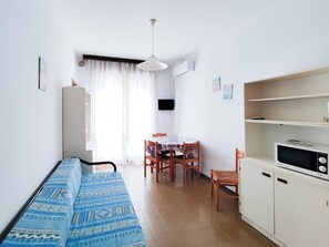 Ein kleiner, heller Raum mit einem Bett, einem Tisch, Stühlen, einer Mikrowelle und einem Schrank.