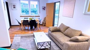 A living room with a sofa, a green chair, a coffee table, and a dining area with chairs and a table. - House with garden & private parking space in Cologne (Köln)
