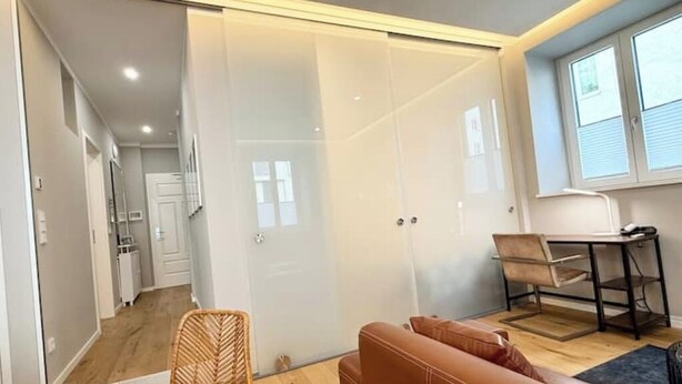 Une pièce moderne dotée d’une porte-fenêtre coulissante donnant sur une cuisine, d’un canapé en cuir marron, d’une petite table avec une chaise et d’une fenêtre avec des stores.