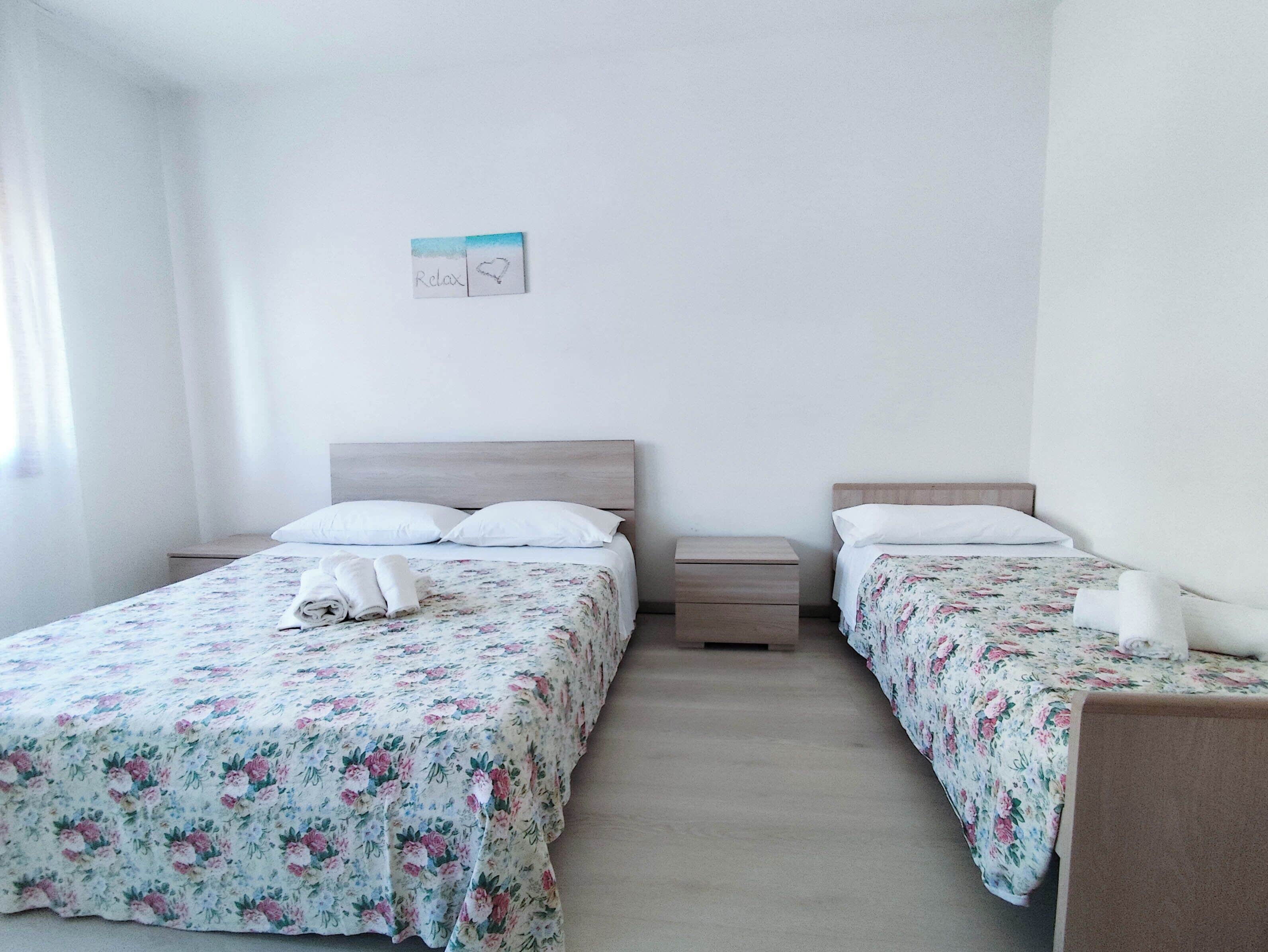 Une chambre double avec deux lits, chacun agrémenté d’une couvre-lit à motifs floraux et de coussins blancs. Il y a une petite table de chevet avec une lampe de chaque côté.
