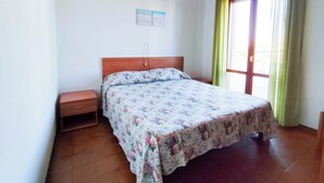 A bedroom with a wooden bed, a nightstand, a small shelf, a window with curtains, and a wall-mounted light fixture. - Spacious Apartment With Balcony by the sea (Jesolo)