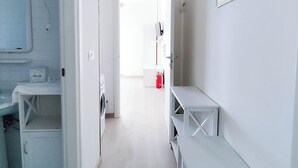 A narrow hallway with a laundry area on the left, a white bench on the right, and a fire extinguisher in the distance. - Spacious Flat With Balcony by the sea (Jesolo)