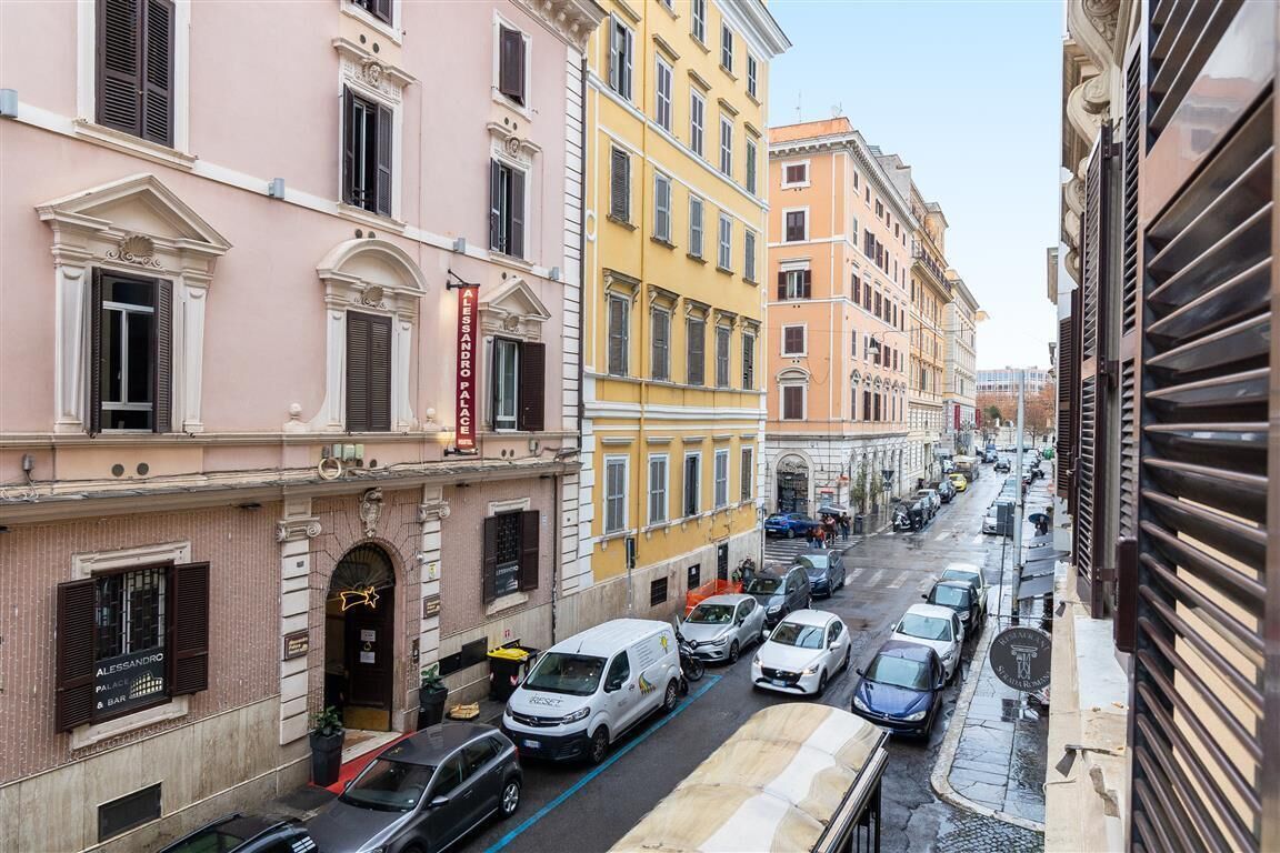 Une vue de rue d’une ville avec des immeubles à plusieurs étages, des voitures garées et un ciel dégagé.