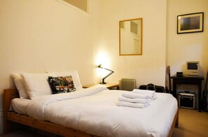 Ein ordentlich bezogenes Bett mit weißen Laken, eine Nachttischlampe, eine Heizung, eine Gitarre und ein Computer auf einem Schreibtisch.