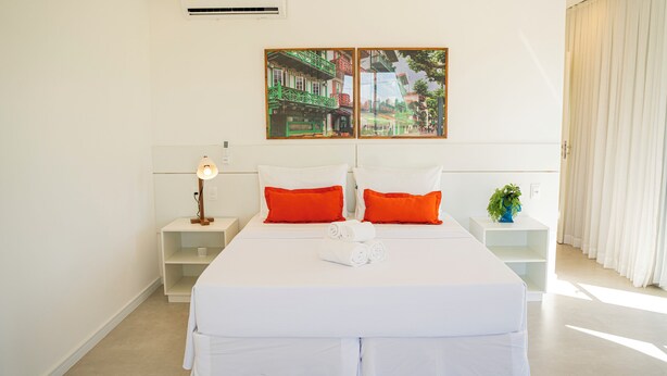A hotel room with a bed, white bedding, two nightstands, a lamp, a potted plant, and a painting of a street scene. - Villa Eivissa Dunes (Trairi)