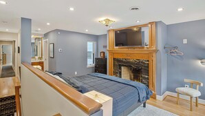 Un dormitorio con una cama, un televisor sobre la chimenea, una silla de madera y un perchero.