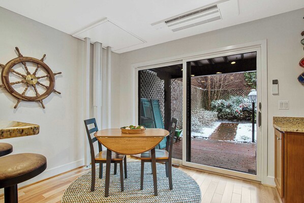 A dining area with a wooden table and chairs, a ship's wheel decoration on the wall, and a view of a snowy garden through a sliding glass door.