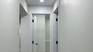 A narrow hallway with white walls and a wooden floor, leading to a room with a door.