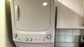 Un appareil compact combinant lave-linge et sèche-linge, doté d’un panneau de commande, installé dans une cuisine avec des meubles blancs et un pare-feu en carrelage.