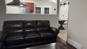 A living room with a black leather sofa, a glass coffee table, and a large mirror reflecting a kitchen area. - Terrific 3-bedroom family-friendly house close to everything in charming McHenry (McHenry)