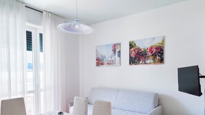A room with a dining table, a sofa, a TV, and two wall-mounted pictures. - Comfy Apartment With Pool Access (Jesolo)
