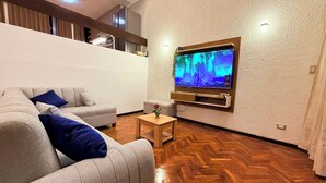 Een moderne woonkamer met een grote flatscreen televisie aan de muur, een grijze bank met blauwe kussens, een klein houten bijzettafeltje en een houten vloer met een herringbone patroon.