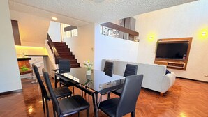 Een moderne woonkamer met een glazen eettafel, zwarte stoelen, een grijze bank en een aan de muur bevestigde televisie.