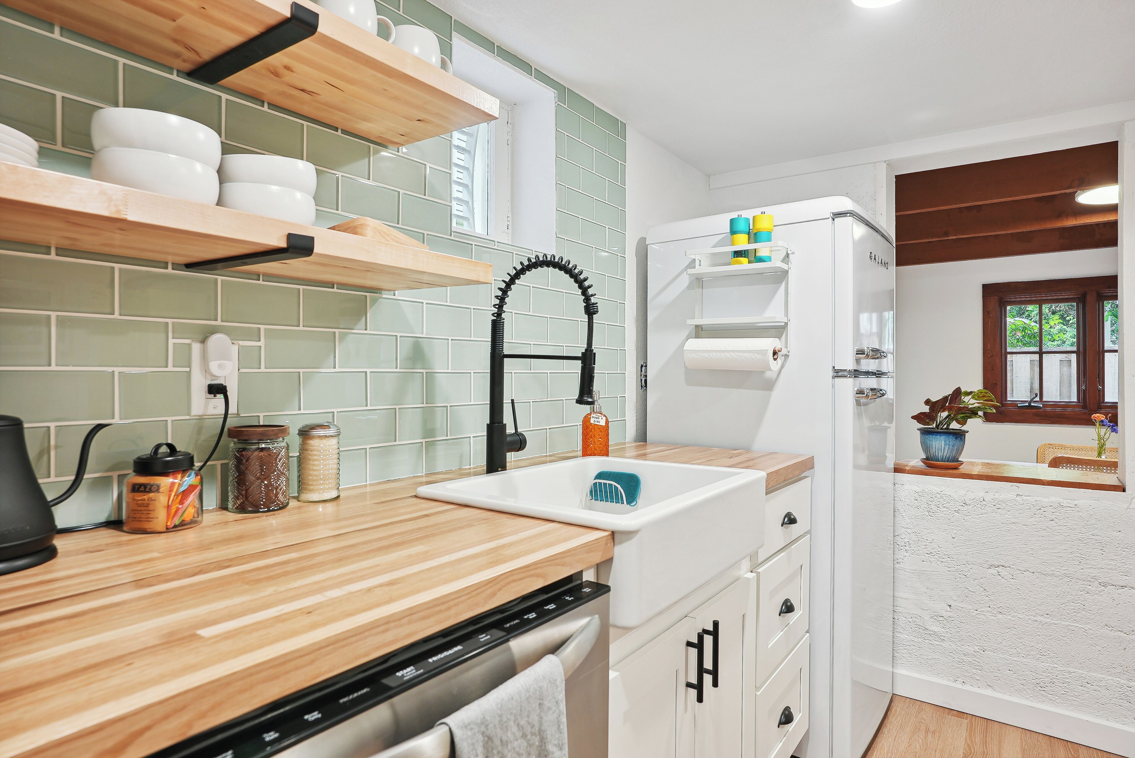A kitchen with wooden countertops, a white sink, and a built-in oven. There are shelves above the counter holding dishes and a refrigerator with a plant on top.