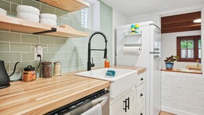 Una cocina con encimeras de madera, un lavamanos blanco y un horno empotrado. Hay estantes sobre la encimera con vajilla y un refrigerador con una planta encima.