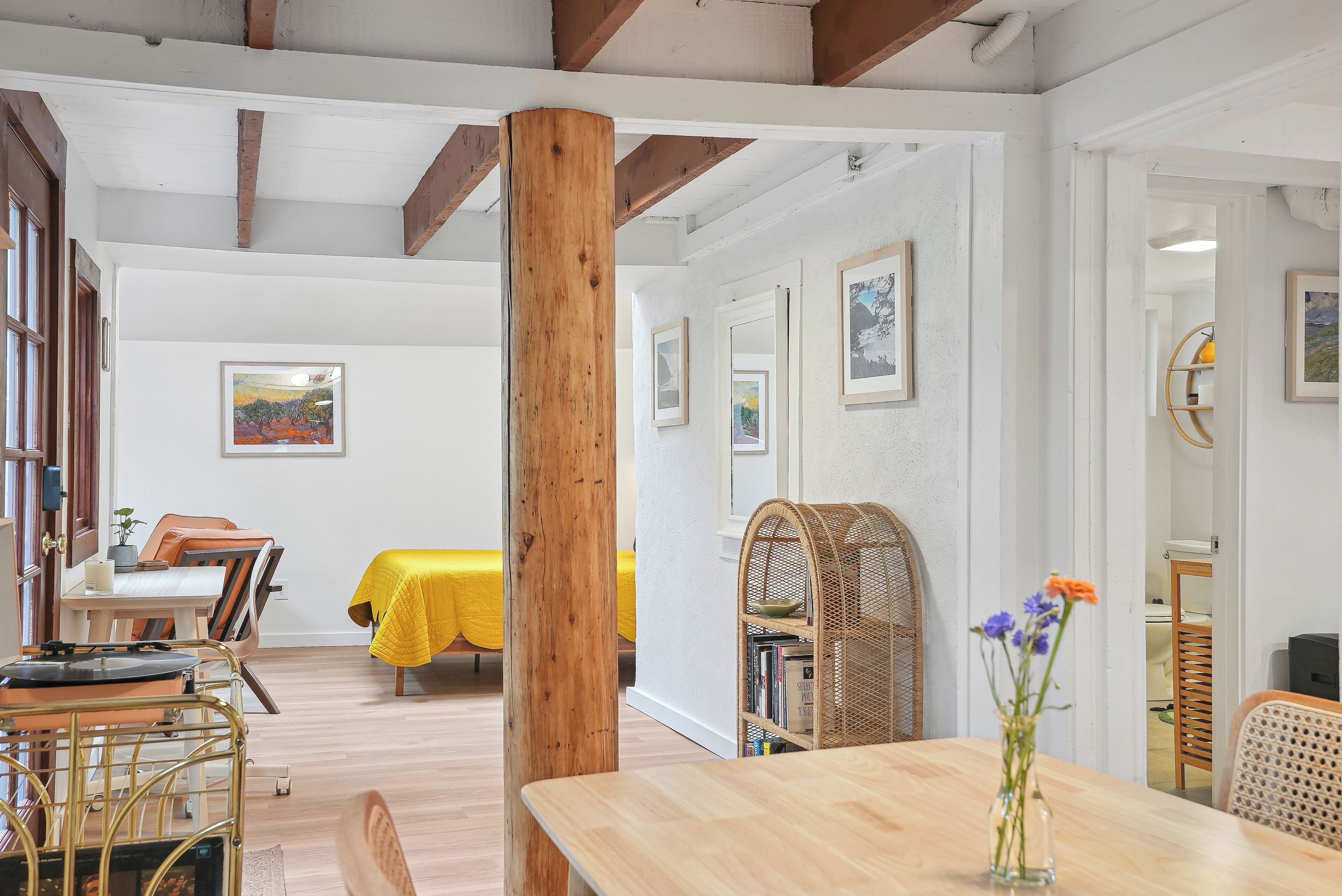 A dining area with a wooden table, chairs, and a yellow tablecloth. There is a bookshelf with books and a vase with flowers.