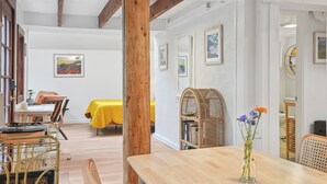 Un comedor con una mesa de madera, sillas y un mantel amarillo. Se observa una estantería con libros y un florero con flores.