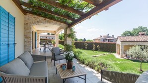 Eine überdachte Terrasse mit Gartenmöbeln, einer Steinwand und Blick auf den umliegenden Garten und die Gebäude.