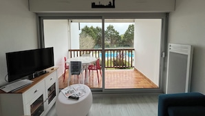 Una sala moderna con un televisor de pantalla plana, un reposapiés blanco con revistas, una mesa de centro de madera y un balcón con vista a una piscina y árboles.