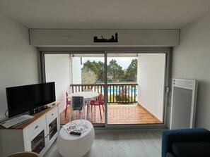 Ein modernes Wohnzimmer mit einem Flachbildfernseher, einem weißen Hocker mit Zeitschriften, einem Holztisch und einem Balkon mit Blick auf einen Swimmingpool und Bäume.