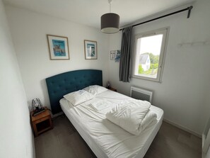 Ein ordentlich gemachtes Bett mit weißen Bettwäsche, einem blauen Kopfteil und einem Nachttisch mit Lampe. An der Wand hängen zwei gerahmte Bilder, und ein Fenster bietet Blick auf ein Haus.