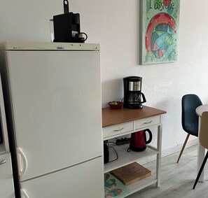 Eine kleine Küche mit einem weißen Kühlschrank, einer Kaffeemaschine auf einem Holztisch und einem roten Wasserkocher.