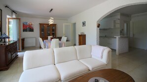 Una sala de estar con un sofá blanco, una mesa de comedor con sillas, un aparador de madera y una zona de cocina con armarios y encimera.