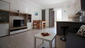Una sala de estar moderna con televisor, mesa de comedor y sillas. Hay una zona de cocina con armarios y un microondas.