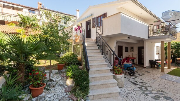 Ein zweistöckiges Haus mit einer Steintreppe, die zu der oberen Etage führt. Es gibt einen Balkon mit Geländer und einem Blumentopf.