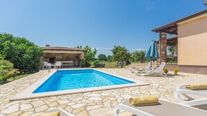 Ein Pool mit Steinplatten, Liegestühlen und einer überdachten Liegefläche mit Tisch und Stühlen.
