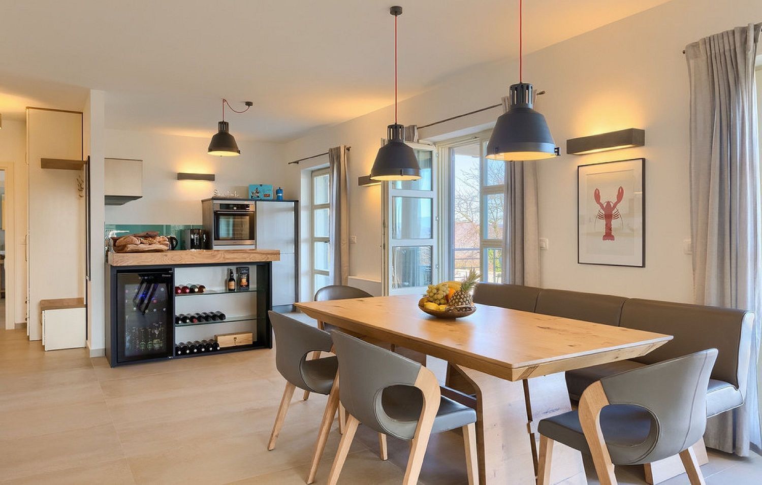 A modern kitchen with a dining area, featuring a wooden table, grey chairs, pendant lights, and a refrigerator.