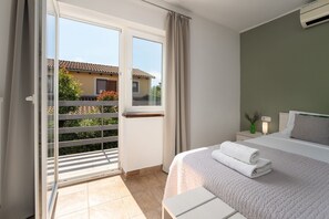 Habitación de hotel con cama, mesita de noche, lámpara y vista a un balcón con barandilla.