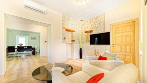 Ein modernes Wohnzimmer mit einer Couch, einem Couchtisch und einem an der Wand befestigten Fernseher. Im Hintergrund ist ein Essbereich mit Tisch und Stühlen zu sehen.