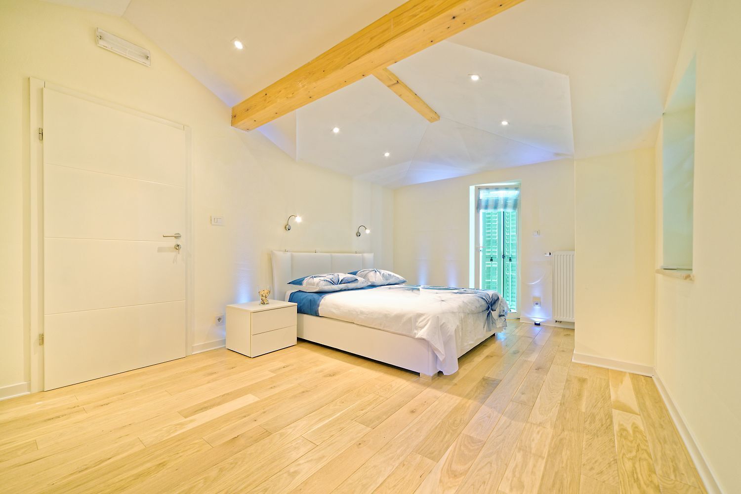 Une chambre avec un plancher en bois, un lit avec une literie blanche, une table de chevet et une fenêtre avec des stores.