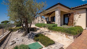 Una casa moderna con facciata in pietra, un portico coperto e un giardino curato con un grande albero e diversi arbusti.