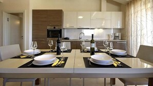 Un'area pranzo moderna con un lungo tavolo apparecchiato per quattro, completa di bicchieri da vino e una bottiglia, in una cucina con mobili bianchi e una parete decorata in legno.
