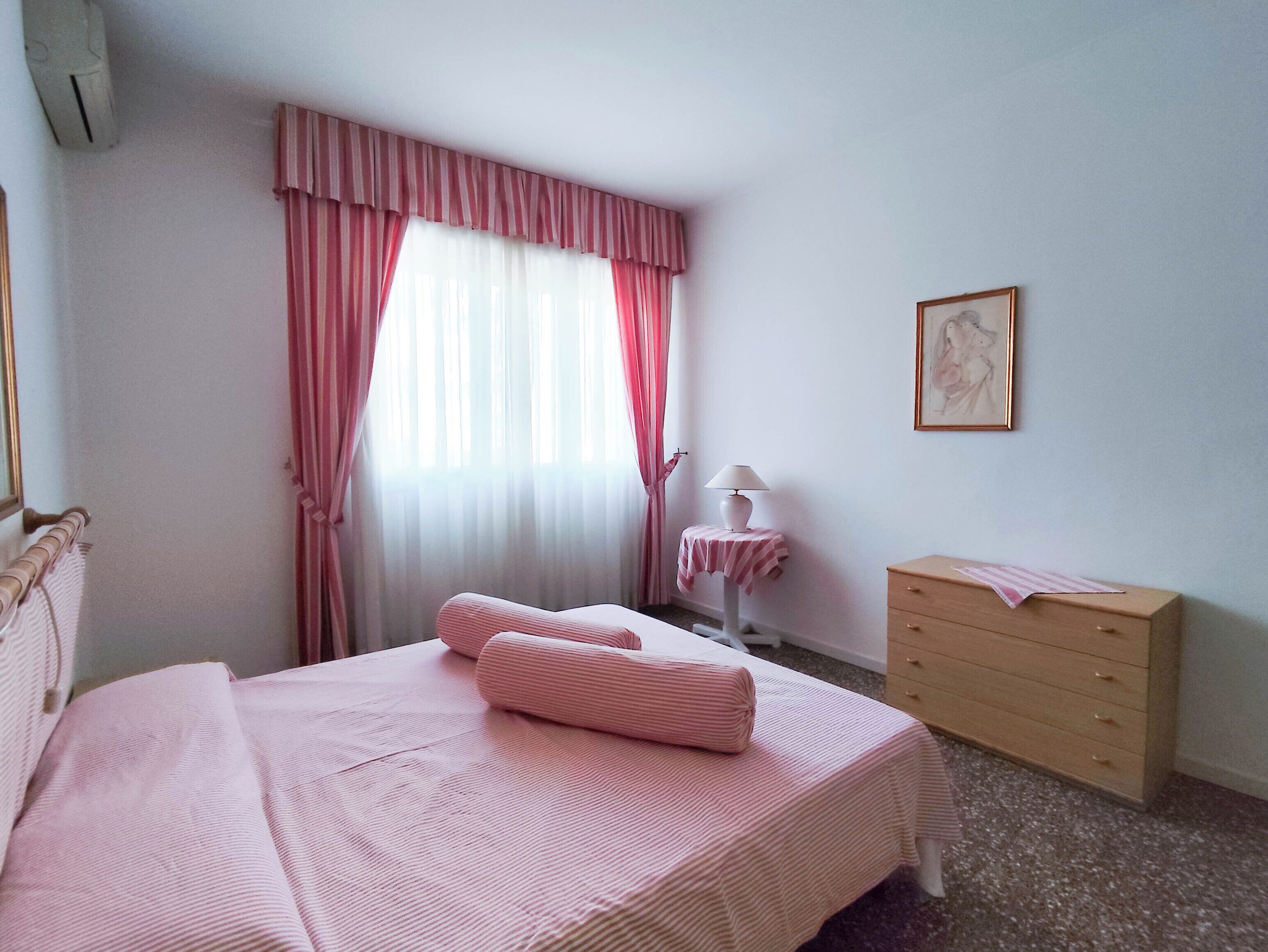 A bedroom with a bed, a dresser, a nightstand, a lamp, and a window with curtains.