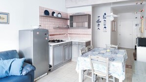 Et kompakt køkken med køleskab, komfur og vask. Der er et spisebord med stole og en blå og hvid dug. En blå sofa står op ad væggen.