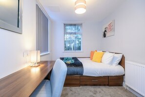 A bedroom with a bed, bedside table, lamp, and a window with blinds.