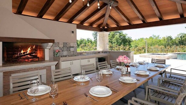 Cocina al aire libre con chimenea de piedra, mesa de comedor de madera dispuesta para seis personas y vistas a la vegetación.
